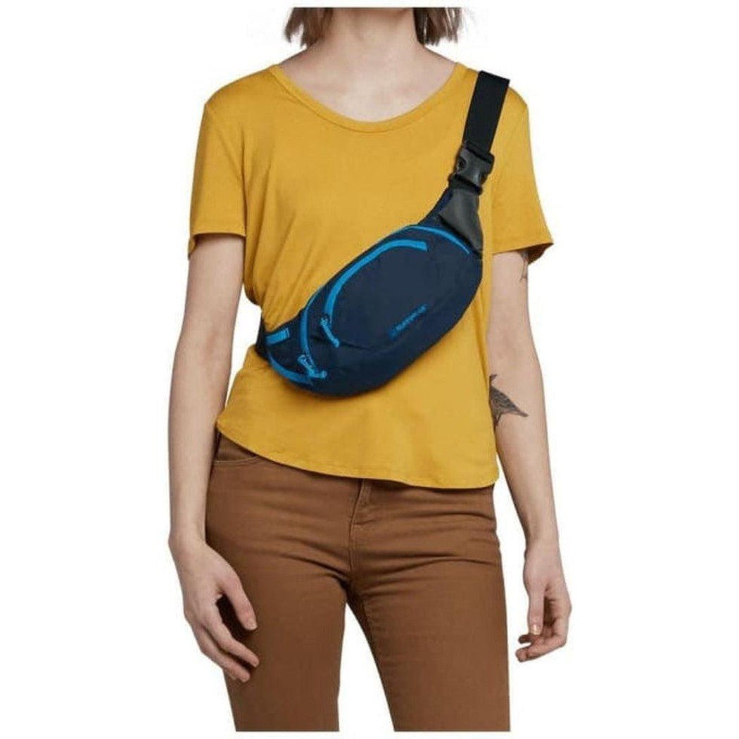 A woman wearing a yellow tee and brown pants models the Ruffwear Home Tail Hip Pack in Aurora Teal from a front angle against a white background.