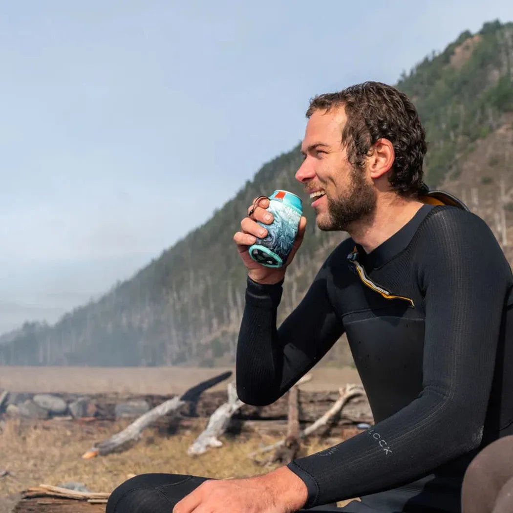 A man in a wetsuit sits outdoors drinking from a Baja Fade Rumpl Bevy Sack against a mountain backdrop.