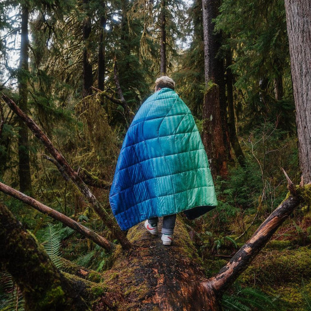 A child wearing the Patch Fade Warm Rumpl Nanoloft Puffy walks on a mossy log in a forest.