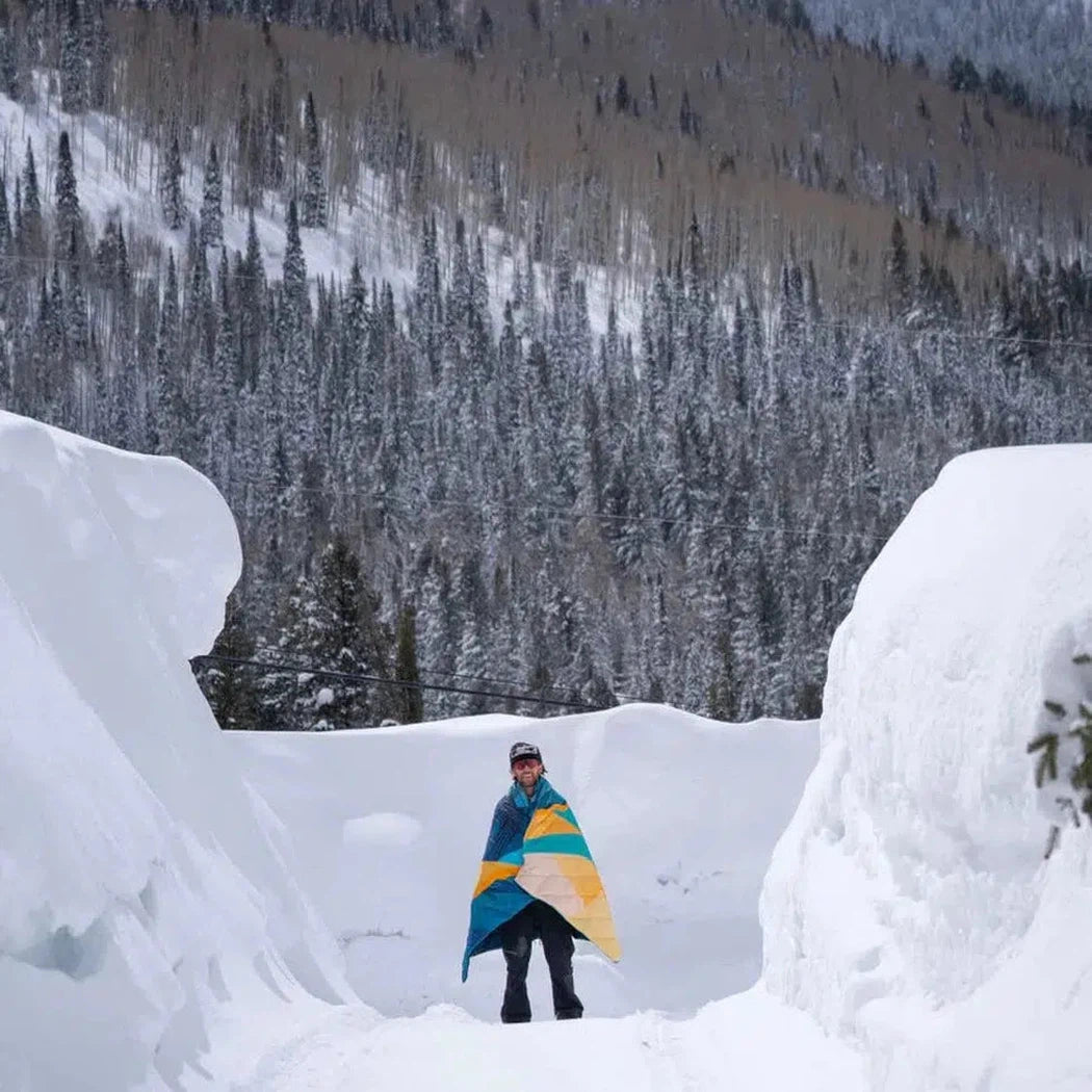 A person stands in a snowy mountain pass wrapped in the Ridgeline Views Rumpl Original Puffy blanket, with snow-covered trees in the background.