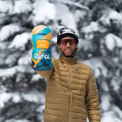A man in a tan jacket holds up a blue and yellow Rumpl Original Puffy blanket in a snowy forest background.