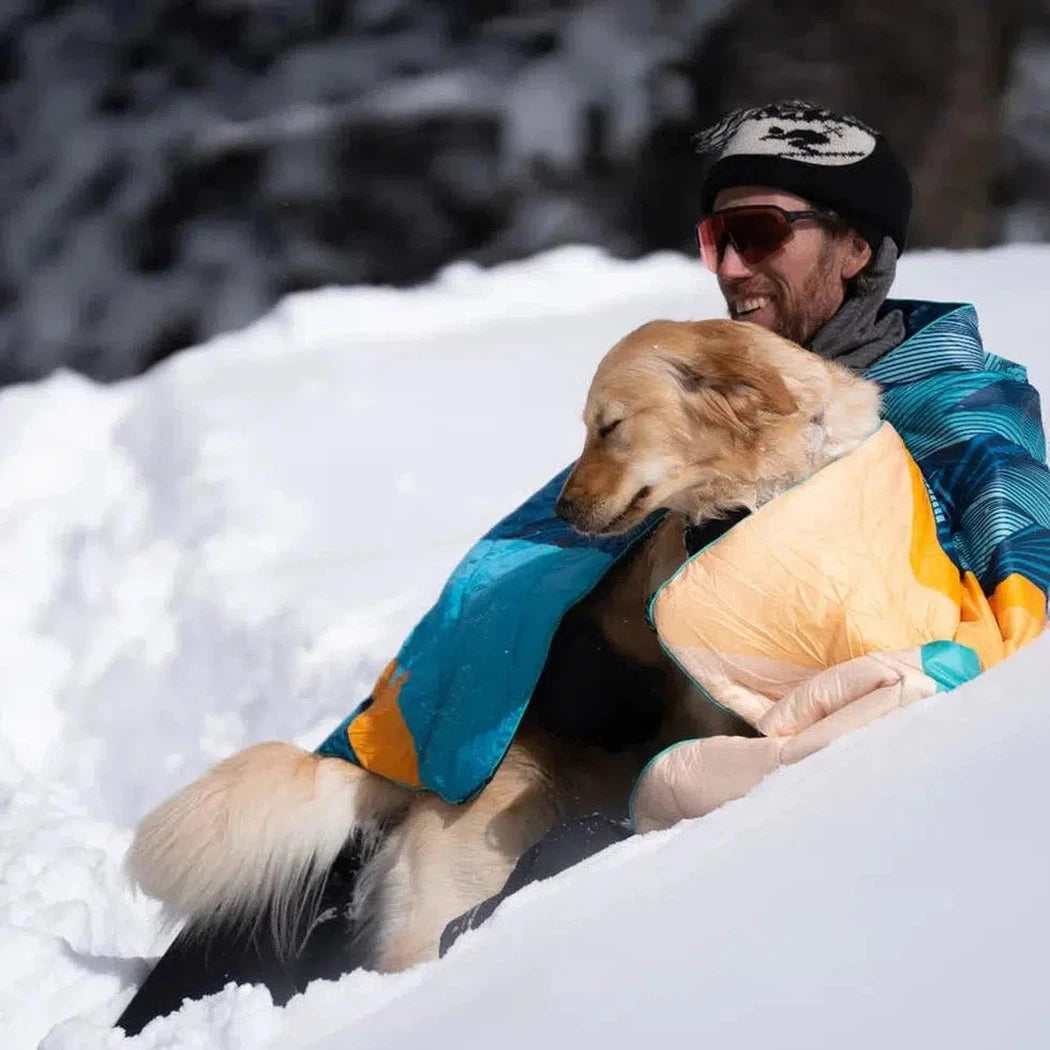 A man and his golden retriever sit in the snow with the Ridgeline Views Rumpl Original Puffy blanket.