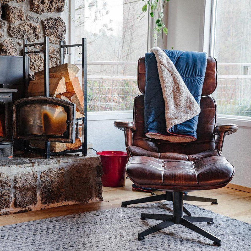 A brown leather chair with a Deepwater Rumpl Sherpa Puffy blanket draped over it sits by a stone fireplace and large window.