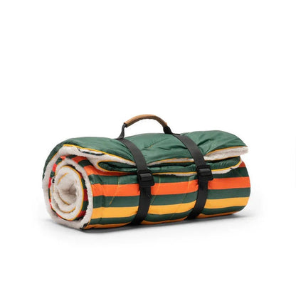 A rolled-up Rumpl Sherpa Puffy blanket with green, orange, and yellow stripes, black straps, and a brown handle against a white background.