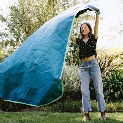 A woman in a black top and jeans holds up the blue Rumpl The Everywhere Mat with green trim in a garden setting.