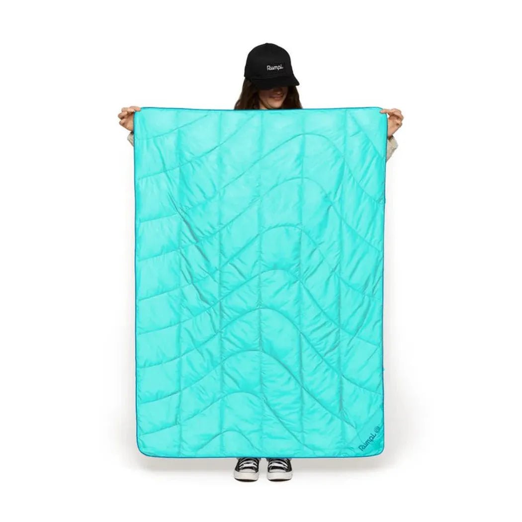 A person wearing a black cap holds up the turquoise Rumpl Travel Puffy blanket against a white background.