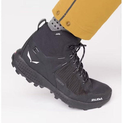 A side view of the black Salewa Women's Pedroc Pro Powertex Mid Boot on a model wearing yellow pants, showing the lugged outsole against a white background.