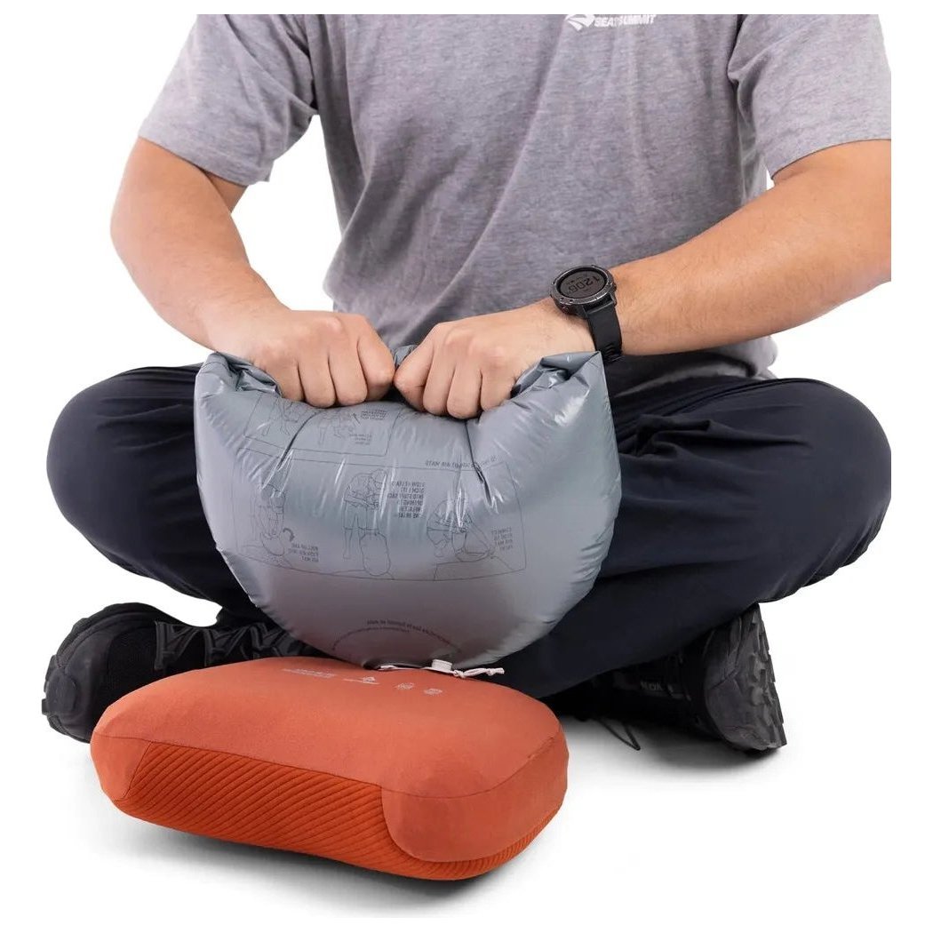 A person sits cross-legged with the Sea To Summit Aeros Premium Pillow in front of a Burnt Olive one, both on a white background.
