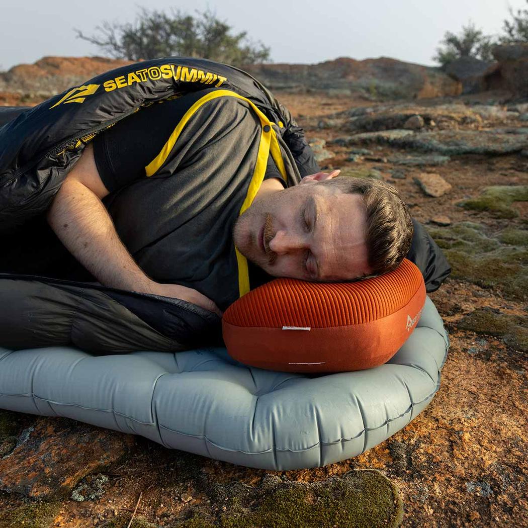 A man sleeps outdoors on a Sea To Summit Aeros Premium Pillow and sleeping pad, with a rocky landscape in the background.