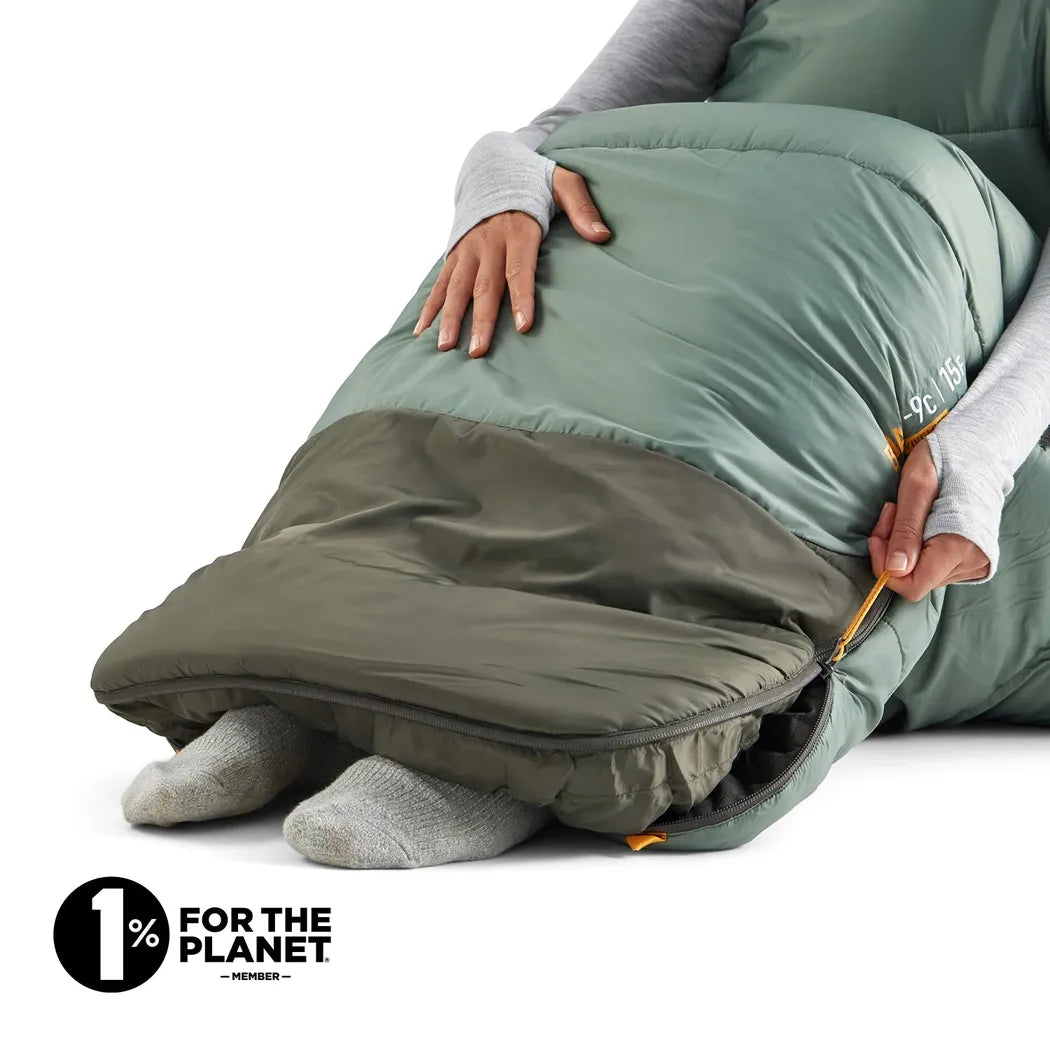 A person wearing a gray long-sleeve shirt and white socks zips the Sea To Summit Boab Synthetic Sleeping Bag 30F|-1C, which is Laurel Wreath Green, against a white background.