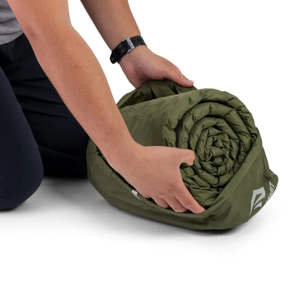 A person kneels while rolling up the olive green Sea To Summit Camp Plus Self Inflating Mat against a white background.