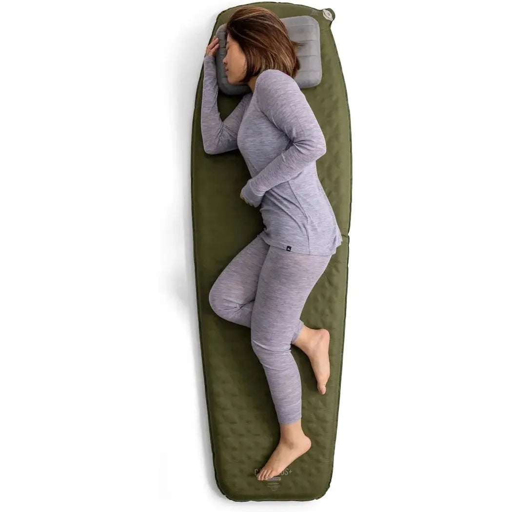 A woman in gray sleepwear lies on a green Sea To Summit Camp Plus Self Inflating Mat with a gray pillow, viewed from above against a white background.