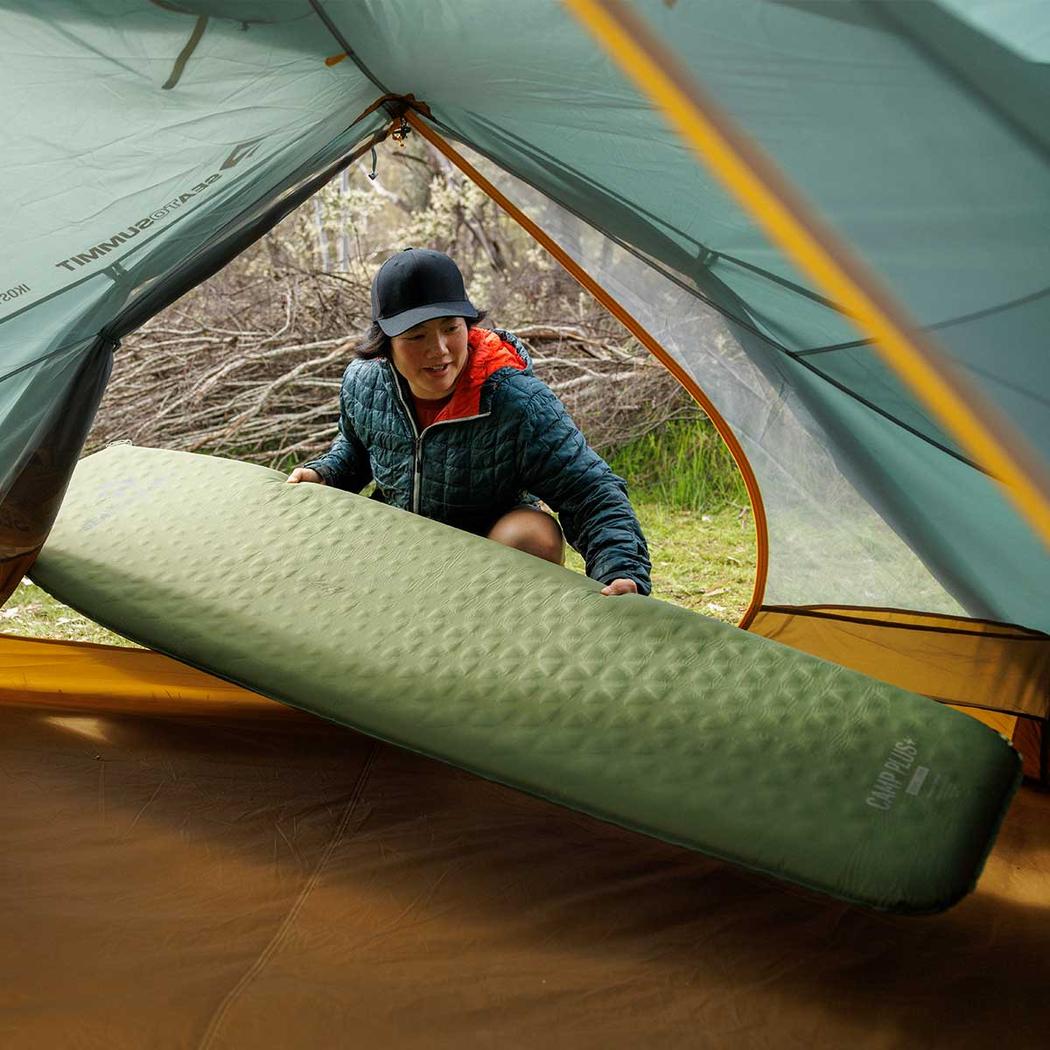 A woman in a blue jacket and black cap places a green Sea To Summit Camp Plus Self Inflating Mat inside a tent with a forest background.