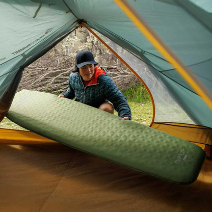 A woman in a blue jacket and black cap places a green Sea To Summit Camp Plus Self Inflating Mat inside a tent with a forest background.