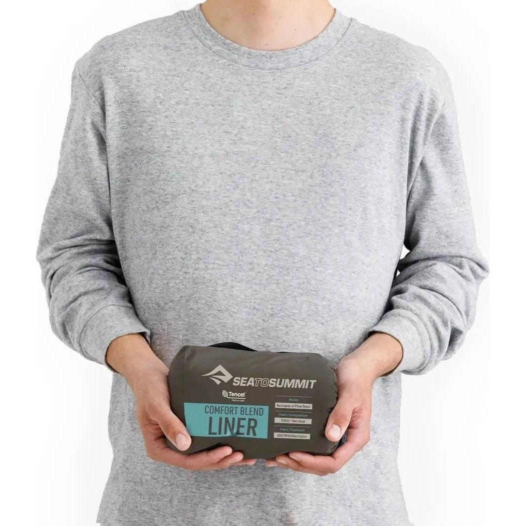 A person wearing a gray sweatshirt holds a Sea To Summit Comfort Blend Liner in their hands against a white background.