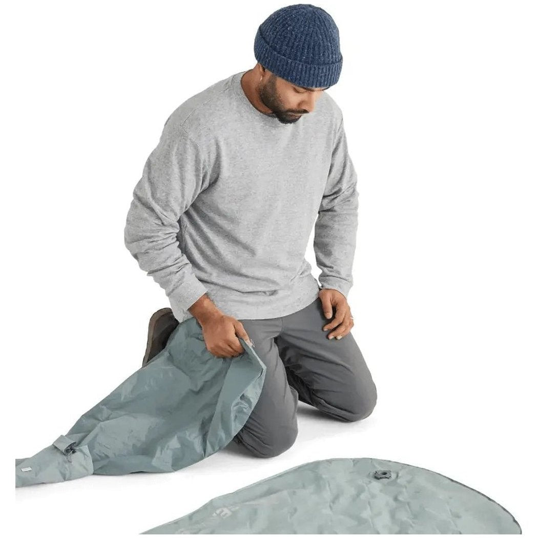 A man kneels while holding the Sea To Summit Ether Light XR Insulated ASC Mat, which is light gray and laid out on a white background.