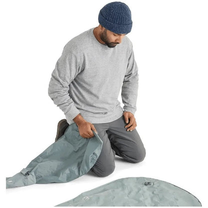 A man kneels while holding the Sea To Summit Ether Light XR Insulated ASC Mat, which is light gray and laid out on a white background.