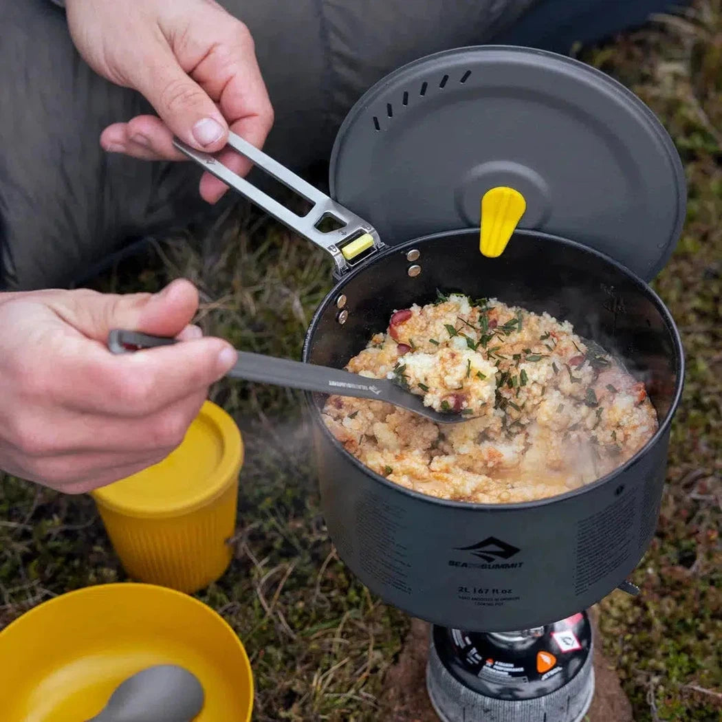 A person uses the Sea To Summit Frontier UL Long Handle Spork to stir food in a charcoal-colored pot on a camp stove outdoors.