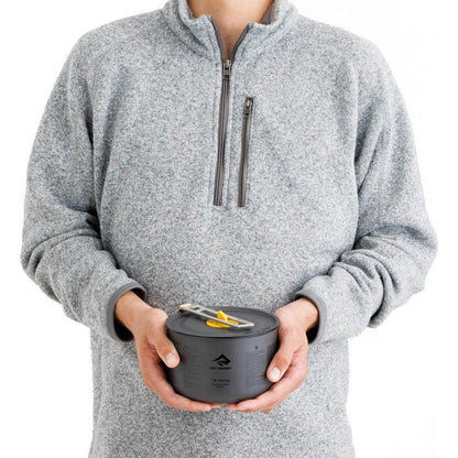 A person wearing a gray fleece holds the Sea To Summit Frontier UL Pot - 1.3L in black, showing the pot and its lid with a yellow and silver tool on top against a white background.