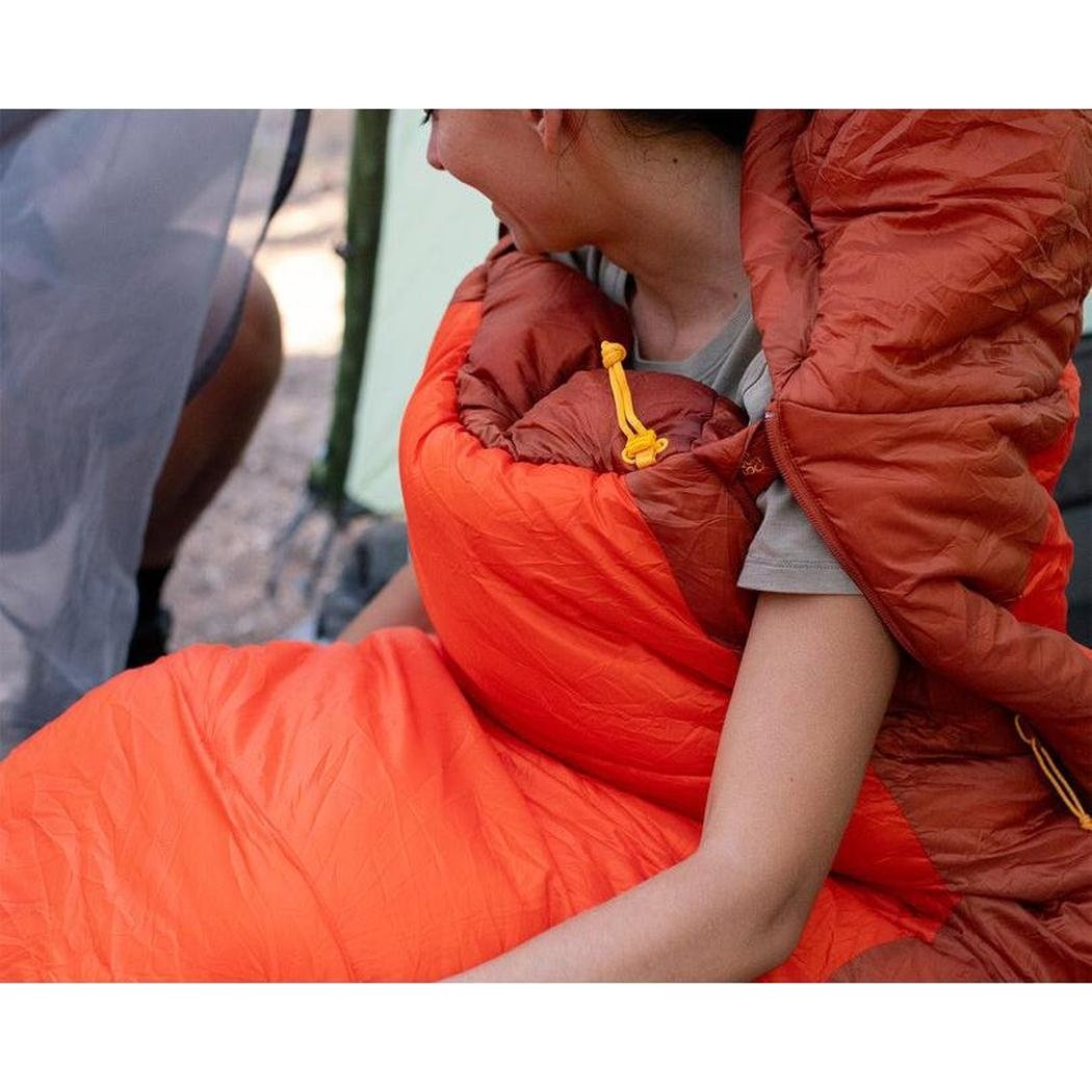 A woman wearing the Sea To Summit Hamelin Women's Synthetic Sleeping Bag in orange sits outside near a tent.