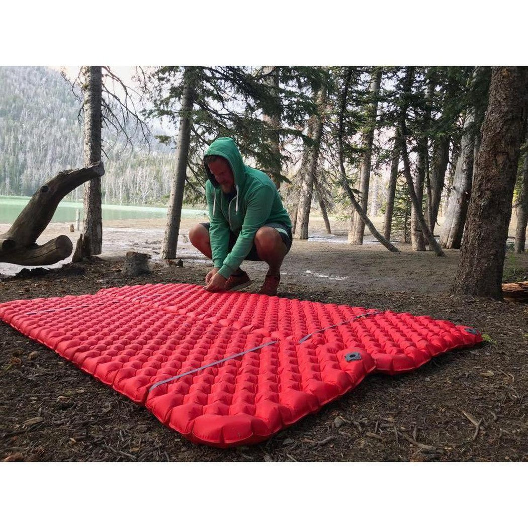 A person in a green hoodie crouches by a red Sea To Summit Mat Coupler Kit Loops on forest ground with trees and a lake in the background.