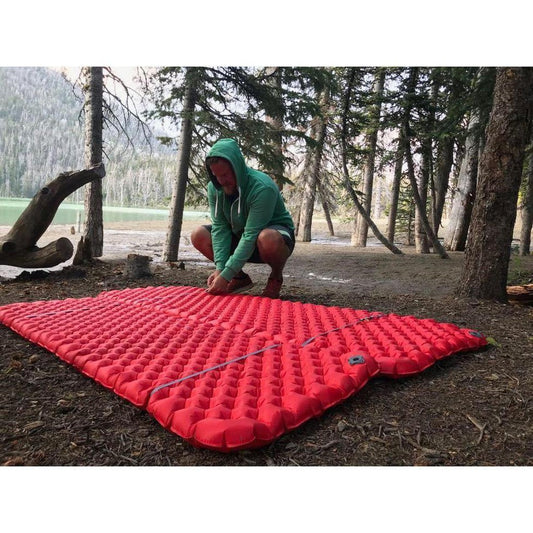 A person in a green hoodie crouches by a red Sea To Summit Mat Coupler Kit Loops on forest ground with trees and a lake in the background.