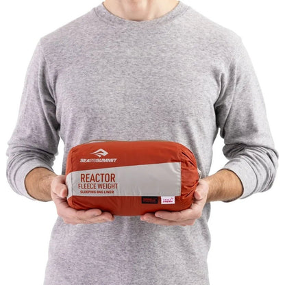 A person in a gray shirt holds a red Sea To Summit Reactor Fleeceweight Liner against a white background.