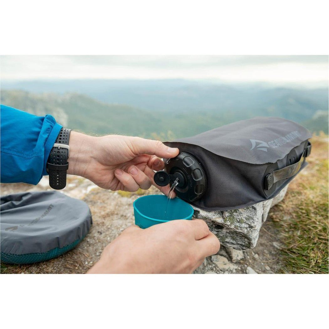 A person pours water from a Sea To Summit Watercell X 4 Liter bag into a teal cup on a rocky mountaintop.