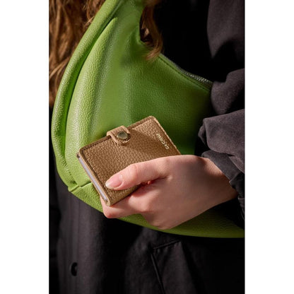 A close-up of the SECRID Miniwallet Pebble in Cappuccino being held by a model wearing a charcoal jacket with a green bag in the background.