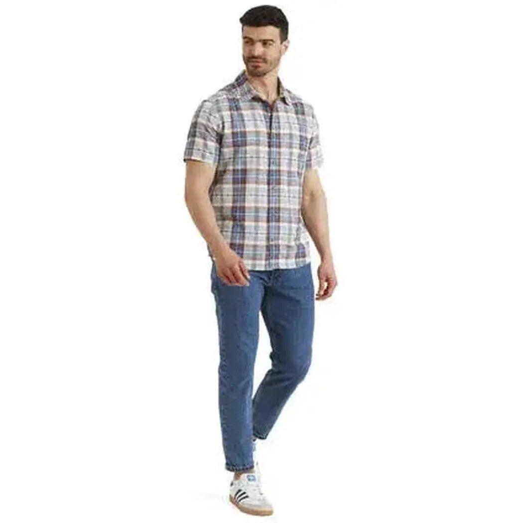 A man wearing the Sherpa Adventure Gear Men's Jampa Plaid Short Sleeve Shirt in Peetho stands against a white background.