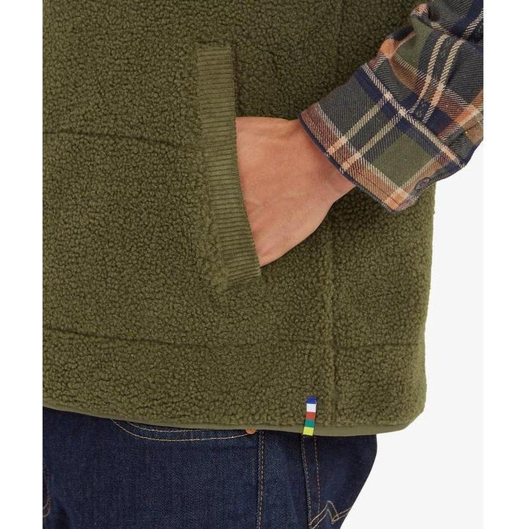 A close-up of the Sherpa Adventure Gear Men's Rangeil Vest in Evergreen, showing a hand in the pocket and a plaid shirt sleeve, against a white background.