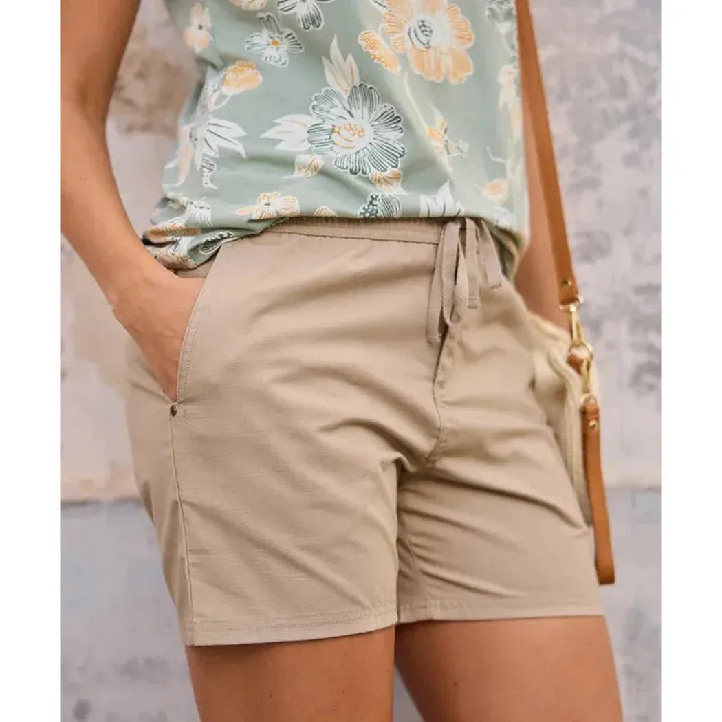 A woman wearing the Sherpa Adventure Gear Sample - Women's Ghoral Short in Bardaya Sand with a floral top and a brown bag, shown from the waist down against a textured wall.