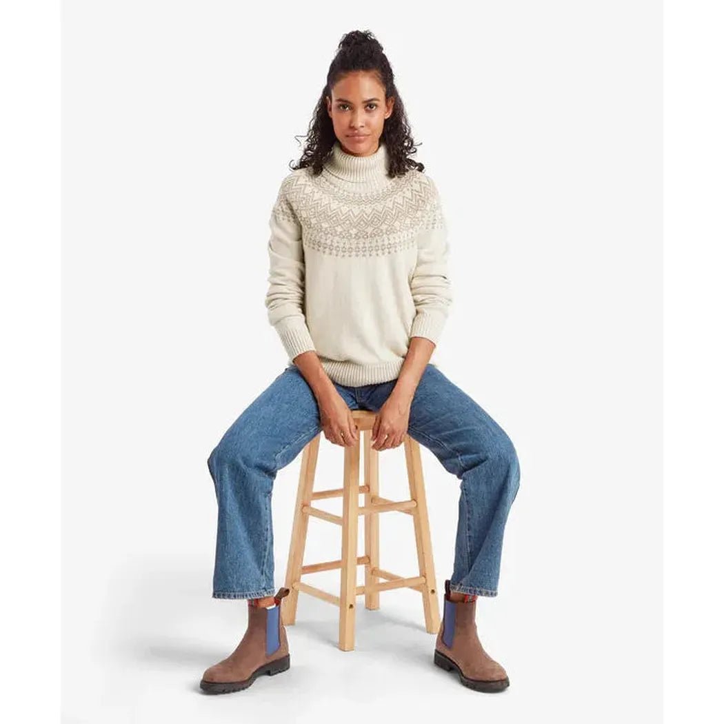 A woman sits on a wooden stool wearing the Sherpa Adventure Gear Samples - Women's Camkilo Roll Neck Sweater in Ecru against a white background.