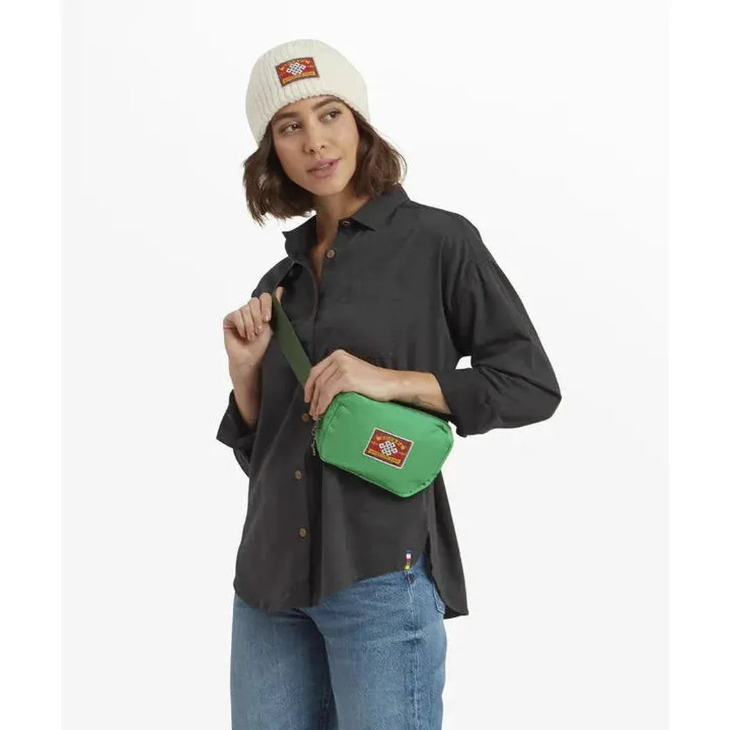 A model wears the Sherpa Adventure Gear Tarcho Belt Bag in Black over a dark shirt and blue jeans against a white background.
