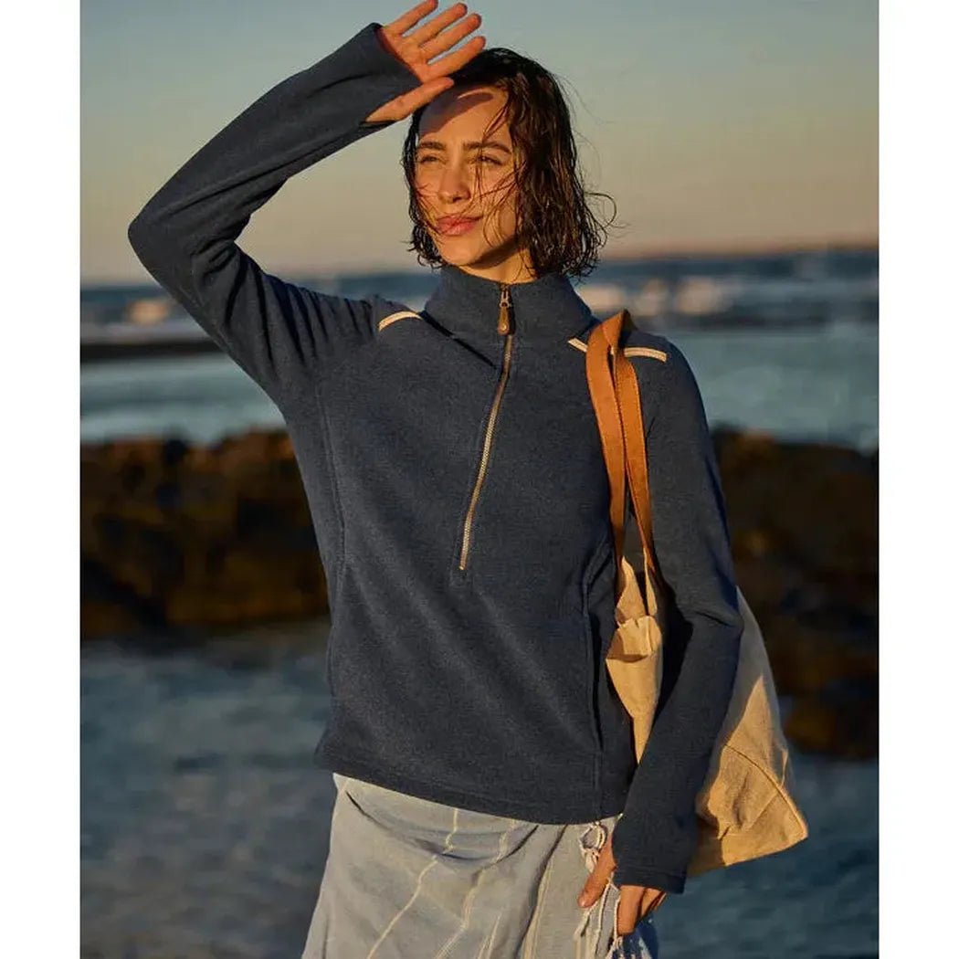 A woman wearing the Sherpa Adventure Gear Women's Rolpa Eco Pullover Zip in Rathee Blue poses by the ocean with a tote bag.