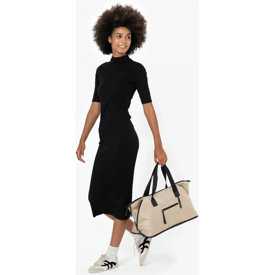 A woman in a black dress and white sneakers holds the Sherpani Eclipse AT bag in Aloe Mist, shown from the side against a white background.