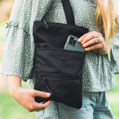 A woman holds the Sherpani Pica Raven crossbody bag, showing its front pocket with a phone and a zippered bottom pocket, against a green outdoor background.