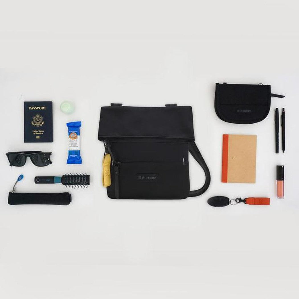 A flat lay of the Sherpani Pica Raven bag with passport, sunglasses, and pens visible on a white background.