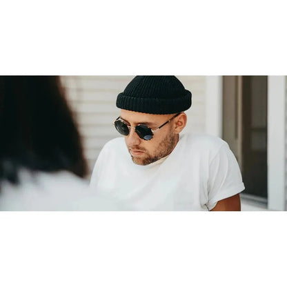 A man wearing the Shwood Hawthorne sunglasses in Black Chrome // Mahogany looks down while wearing a black beanie and white tee in an outdoor setting.
