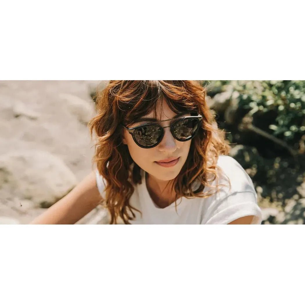 A woman wearing Shwood Kinsrow sunglasses poses outdoors with a rocky background.