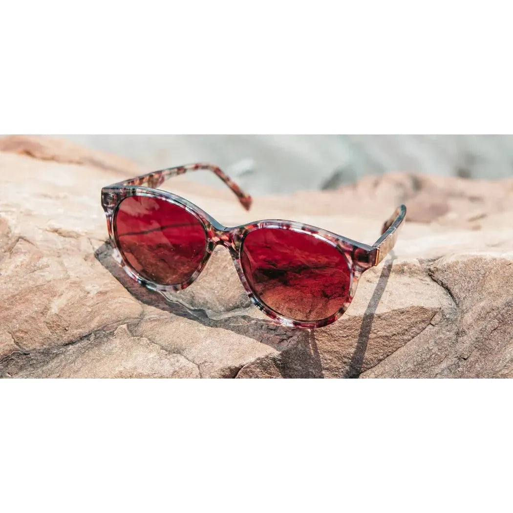 Side view of the Shwood Madison sunglasses with red lenses resting on a rocky surface.
