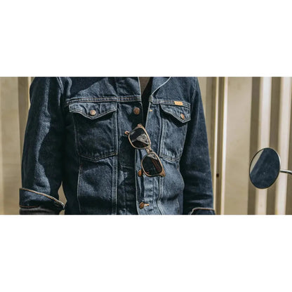 A close-up of a person wearing a dark blue denim jacket with a pair of Shwood Prescott sunglasses hanging from the front pocket, against a light-colored background.