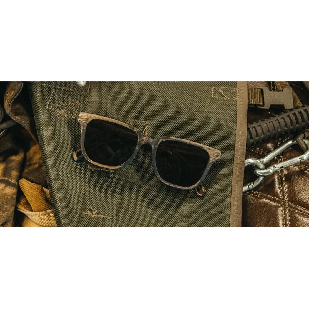 A pair of Shwood Prescott sunglasses in Distressed Dark Walnut rests on a green fabric surface with metal hardware visible nearby.