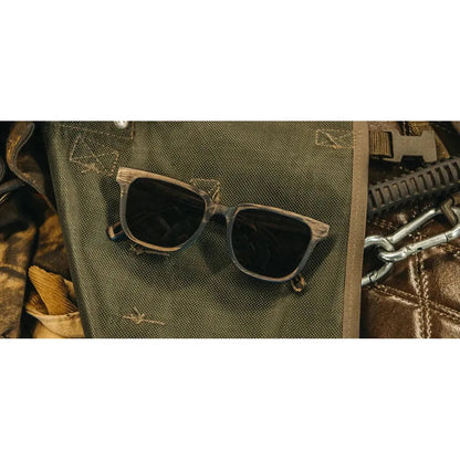 A pair of Shwood Prescott sunglasses in Distressed Dark Walnut rests on a green fabric surface with metal hardware visible nearby.