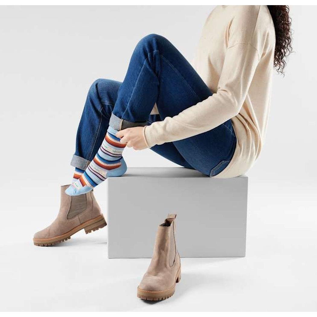 A woman in blue jeans and a cream sweater sits on a gray block, adjusting her colorful striped Smartwool Everyday Margarita Crew socks while wearing tan boots, with one boot on the floor below.