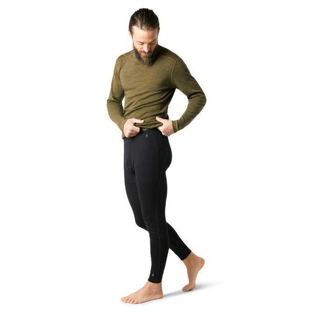A man wearing black Smartwool Men's Classic Thermal Merino Base Layer Bottoms stands barefoot against a white background.