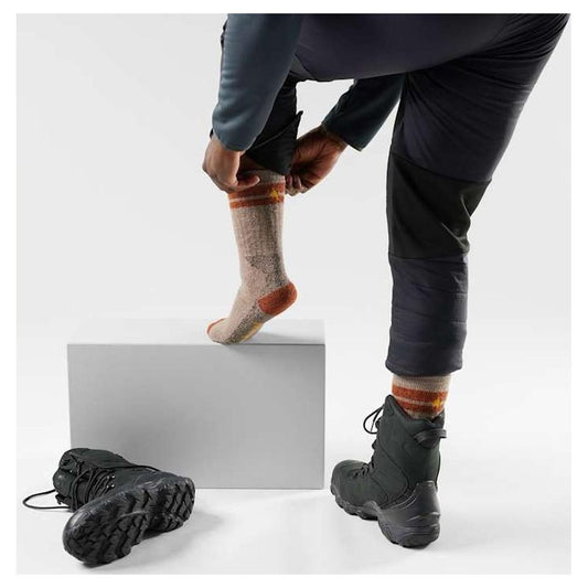 A person adjusts a pair of Fossil Smartwool Mountaineer Maximum Cushion Tall Crew Socks while wearing Charcoal/Graphite boots, with a white background and a gray block.