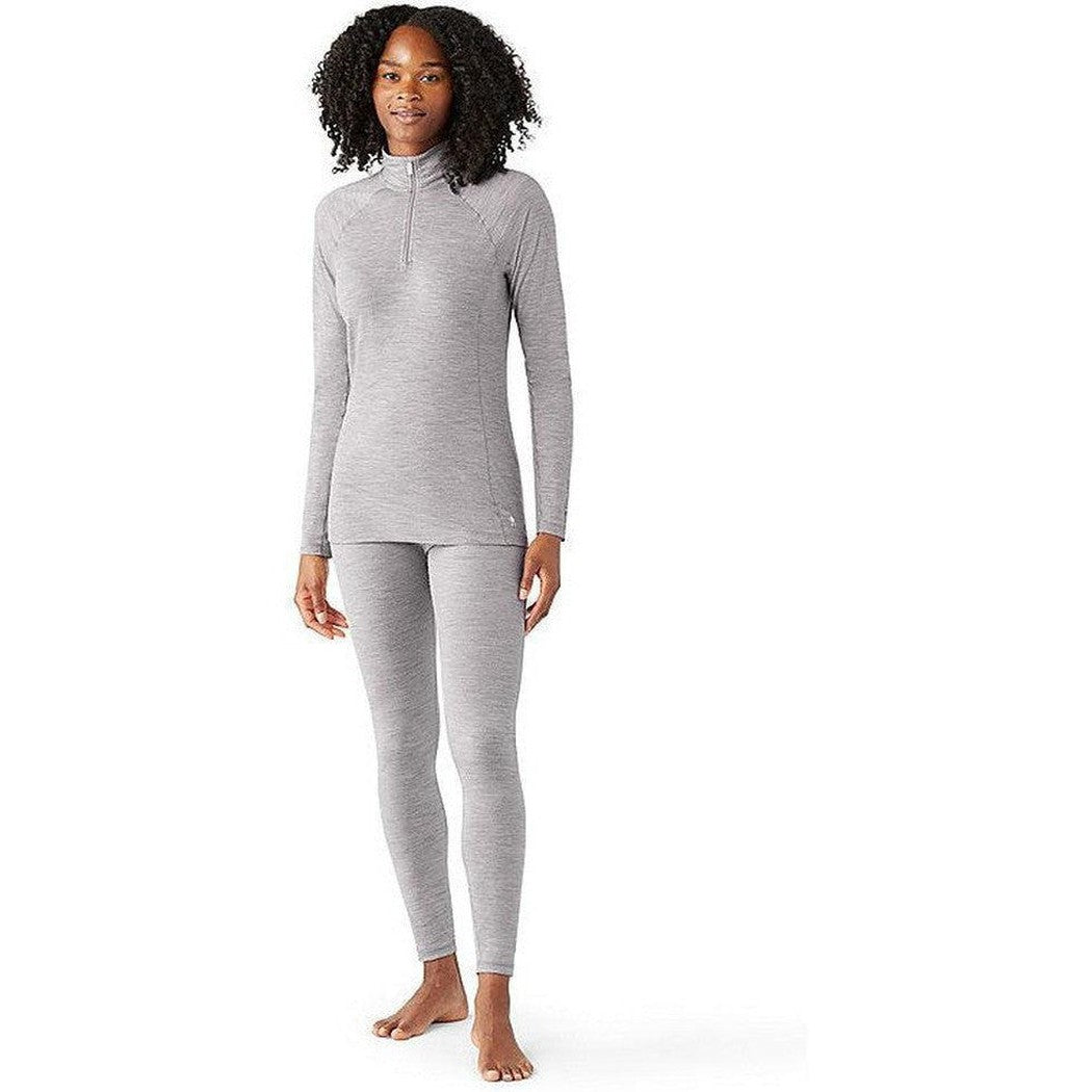 A woman wearing the Smartwool Women's Classic All-Season Merino Base Layer Bottom in Light Gray Heather stands barefoot against a white background.
