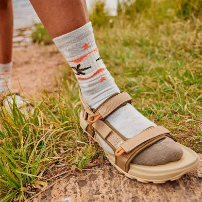 A close-up of a woman wearing Smartwool Women's Hike Light Cushion Otter Float Crew Socks in Ash with a sandal and grassy background.