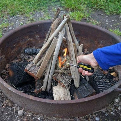 A hand uses the Soto Outdoors Pocket Torch XT to light a campfire in a metal ring on a grassy patch.